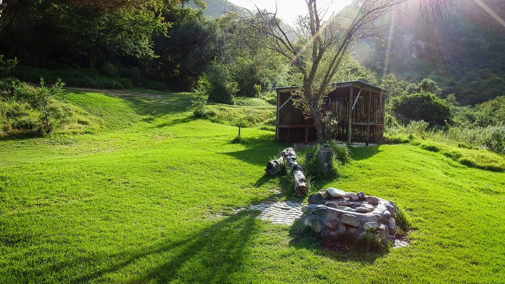 Lush green lawn campsite at Bruintjieskraal featuring a stone braai place and wooden undercover utility structure in a sunny valley